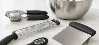 Box grater, garlic press and bench scraper beside a stainless steel mixing bowl, practical everyday kitchen tools